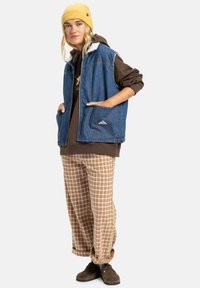 Ungdom som har på seg en gul lue, blå denimvest med fleecefôr, brun hettegenser, rutete bukser og brune slip-on sko, står avslappet.