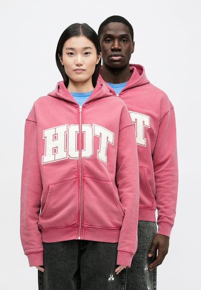 Pink zip-up hoodie with "HOT" in large cream letters, featuring a drawstring hood, front pockets, and ribbed cuffs. Worn over blue shirts.