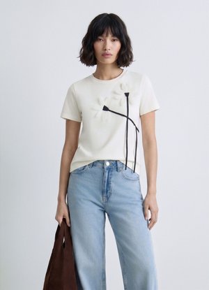 Donna con indosso una t-shirt bianca con motivi floreali neri abbinata a jeans azzurri, che tiene una borsa marrone su uno sfondo chiaro e semplice.