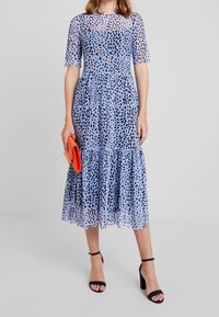 Světle modré šaty s černým a oranžovým tečkovaným vzorem, průhlednými krátkými rukávy, volánkovým spodním lemem a přiléhajícím pasem. Doplněné oranžovou kabelkou.