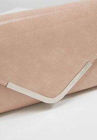 Svetle béžová kabelka na ruku s hladkým povrchem, se stříbrným kovovým detail na rohu a jemným gravírováním loga.