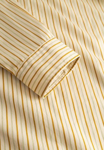 Keltainen ja kerma raidallinen kangas, jossa ohuita vihreitä viivoja. Lähikuva huolellisesti käännetystä hihansuun kalvosta, joka esittelee kankaan tekstuurin ja kuvion.