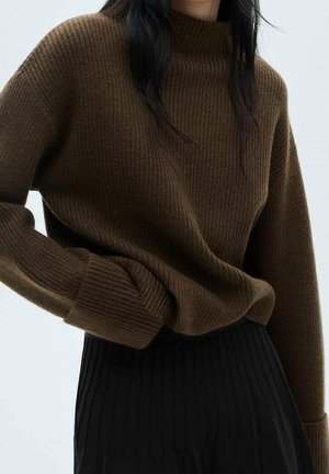 Femme portant un pull côtelé marron à manches longues, rentré dans un pantalon noir plissé taille haute, sur un fond uni.
