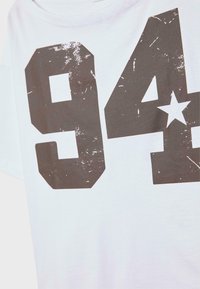 Valkoinen puuvillapaita, jossa on suuri, kulunut tummanharmaa painatus numeroista "94" ja tähdestä. Paidassa on pyöreä kaula-aukko ja lyhyet hihat.