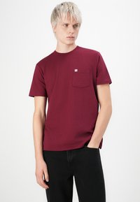 Mørkerød bomulds t-shirt med korte ærmer, rund hals og en brystlomme. Har et lille mærke på lommeområdet.