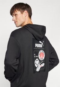 Felpa grigia scura con cappuccio caratterizzata da una stampa grafica sul retro, che mostra il logo Puma e l'emblema del FC St. Pauli con elementi calcistici.