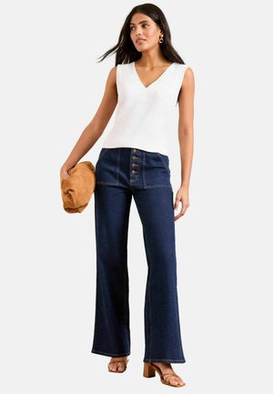 Naine kannab valget varrukateta V-kaelusega topi, tumedat sinist kõrge vöökohaga laia säärtega teksapükse nuppudega, beežikaid kontsakingi, hoides pruuni käekotti.