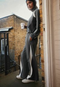 Gray athletic tracksuit with white side stripes, featuring a high collar and elastic cuffs, paired with white sneakers, on a textured surface.