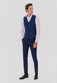 Granatowa kamizelka z pięcioma guzikami na jasnoróżowej koszuli, zestawiona ze skrojonymi granatowymi spodniami w kratę i czarnymi skórzanymi butami.