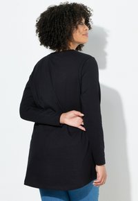 Zwarte longslavige shirt met een ontspannen pasvorm, afgeronde zoom en zachte katoenen stof, gecombineerd met een blauwe spijkerbroek. Het achteraanzicht benadrukt de silhouet.
