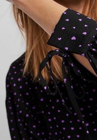Černá látková blůza s fialovým srdíčkovým vzorem, s nabíranými rukávy a zavazovacími manžetami. Texturovaný a lehký materiál.