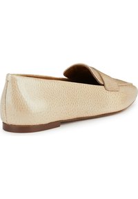 Zapato slip-on de cuero beige con superficie texturizada y suela plana. Presenta una solapa decorativa en la parte superior. Diseño de punta redondeada.
