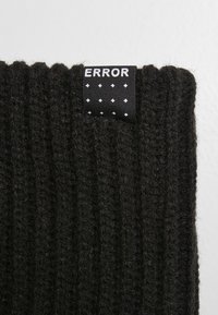 Căciulă neagră din tricot cu model în relief. Dispune de o etichetă țesută cu cuvântul "EROARE" în alb pe un fundal negru cu puncte.