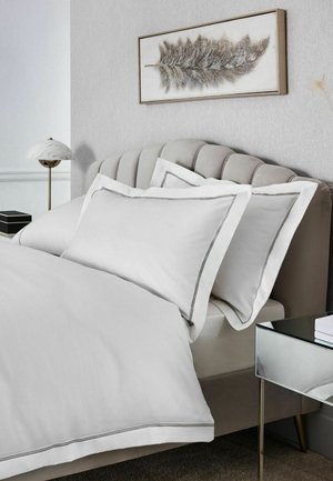 Bett mit gepolstertem Kopfteil, weißen Kissen und Bettdecke mit grauem Rand, Nachttisch mit Lampe und Federkunst an hellgrauer Wand.