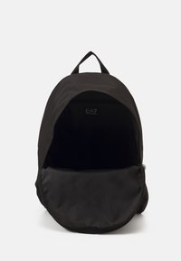 Rucsac din țesătură neagră, cu vârf rotunjit, deschidere cu un singur fermoar și un buzunar interior. Textură netedă, cu detalii hardware minime.