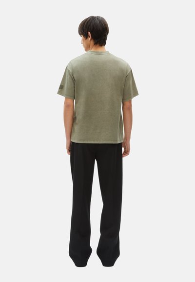 Camiseta verde oliva de manga corta con un corte relajado, escote redondo y costuras visibles, combinada con pantalones negros de pierna ancha. Vista trasera.