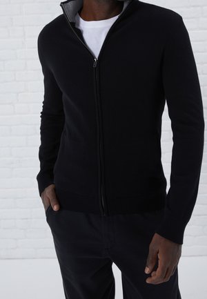 Maglione zip-up nero in tessuto knit con colletto alto e fodera interna grigia. Presenta polsini e orlo a costine, indossato su una camicia bianca.