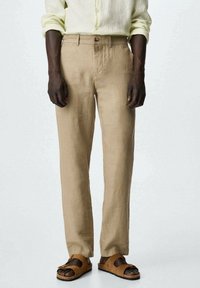 Pantalon en lin beige à coupe droite, avec une fermeture à bouton et des passants pour ceinture. Associé à des sandales marron à enfiler.