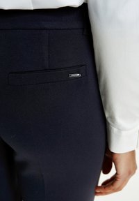 Marina byxor med en slät yta och en bakficka med en silverfärgad "TAIFUN"-tag; tillsammans med en vit långärmad skjorta.