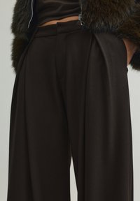 Pantaloni in tessuto marrone scuro, caratterizzati da vita alta con pieghe e silhouette sartoriale. Texture liscia e linee pulite.