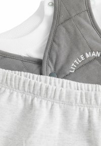 Salopette en coton gris avec un design matelassé et un débardeur blanc en dessous. Elle dispose de boutons pression et d'une taille élastique. Texte "PETIT HOMME" brodé.