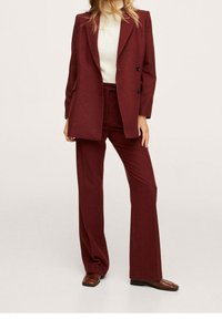 Costume sur mesure couleur bordeaux avec un blazer à double boutonnage et un pantalon à jambes larges. Tissu texturé et design minimaliste. Des chaussures marron complètent la tenue.