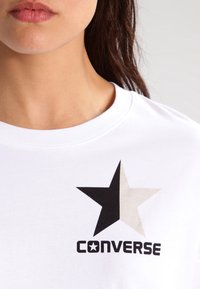 Biała bawełniana koszulka z grafiką czarno-beżowej gwiazdy oraz wyboldowanym napisem "CONVERSE" w czerni. Okrągły dekolt z krótkimi rękawami.