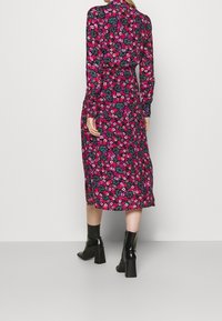 Frau steht mit dem Rücken zur Kamera und trägt ein langärmliges, mittellanges Blumenkleid mit pinken, roten und lila Blumen, kombiniert mit schwarzen, hohen Stiefeletten.