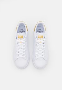 Par av vita Adidas Stan Smith-sneakers med beige hälaccenter och orange logotags, sedda från ovan mot en vit bakgrund.