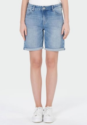Short en jean bleu clair à ourlet roulotté porté avec des baskets blanches, jambes visibles de mi-cuisse aux pieds sur un fond uni.
