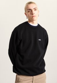 Black crewneck sweatshirt made of soft fabric, featuring the Vans logo in white on the chest and relaxed fit. Worn with beige pants.