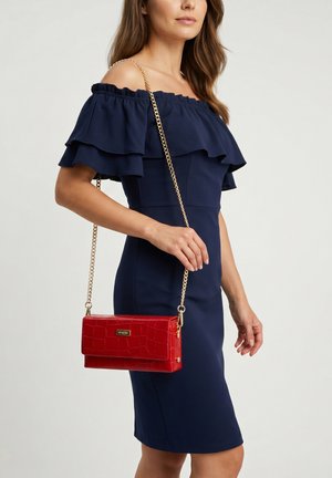 Kvinde iført navy blå off-shoulder kjole, der holder en rød håndtaske med krokodilletekstur og guldkæde skulderrem over skulderen.