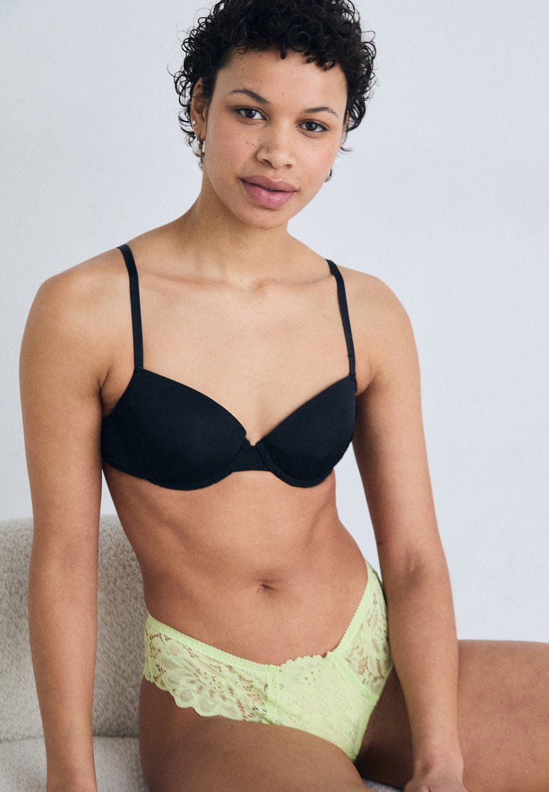 Sotto una tonalità neutra, un reggiseno nero con pizzi e una mutandina in pizzo giallo neon, caratterizzati da spalline regolabili e intricate trame.