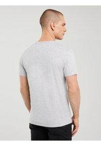Mand med kort hår iført en simpel lysegrå T-shirt med korte ærmer og sorte jeans, stående med ryggen mod kameraet foran en ensfarvet baggrund.