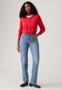 Cardigan rouge avec fermeture à boutons, chemise blanche en dessous, associé à un jean droit bleu clair et des ballerines noires, le tout sur fond neutre.