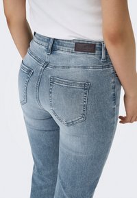 Femme portant un jean denim bleu clair et un haut blanc, vue de dos avec poches visibles et une étiquette de marque en cuir sur la ceinture.