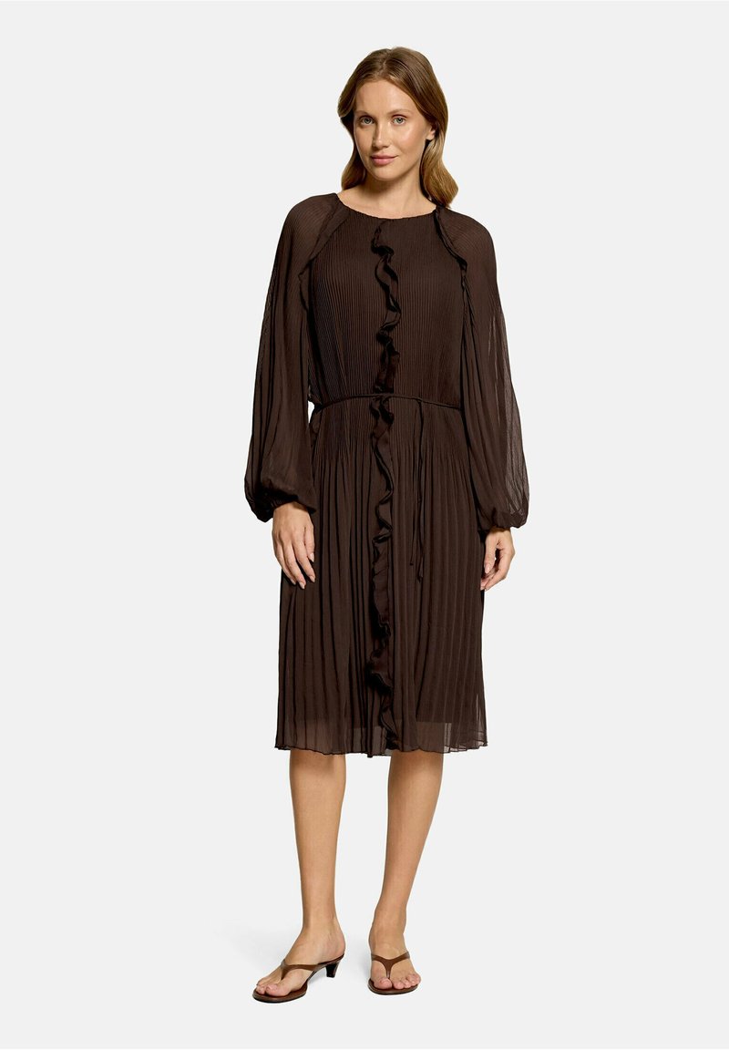 Femme debout portant une robe plissée marron foncé à manches longues avec des volants sur le devant et des sandales ouvertes à talons moyens.