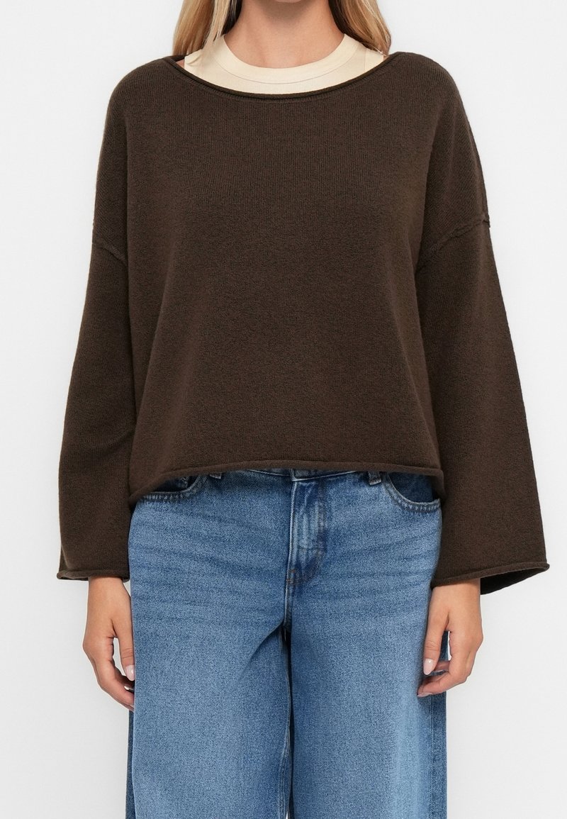Femme portant un pull en maille lâche marron foncé sur un haut beige avec un jean large bleu clair sur un fond uni.