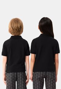 Dwoje dzieci w czarnych koszulkach polo z widocznymi kołnierzykami i podwiniętymi rękawami, stojących plecami do siebie, w wzorzystych czarno-białych spodniach.