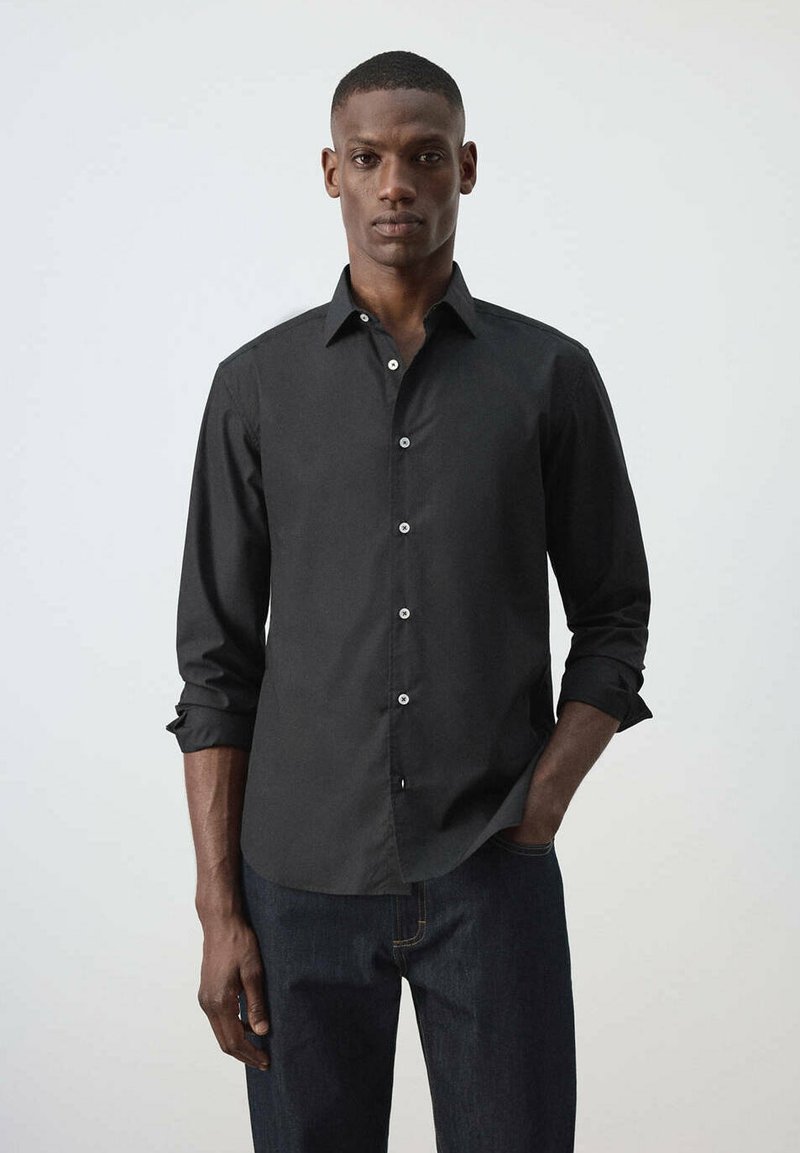 Homme portant une chemise noire à manches longues boutonnée et un jean foncé, debout avec une main dans la poche devant un fond clair uni.