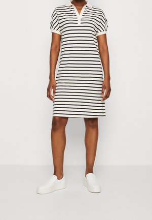 Kvinde iført en kortærmet kjole med krave og sorte og hvide vandrette striber samt hvide slip-on sko, stående foran en hvid baggrund.