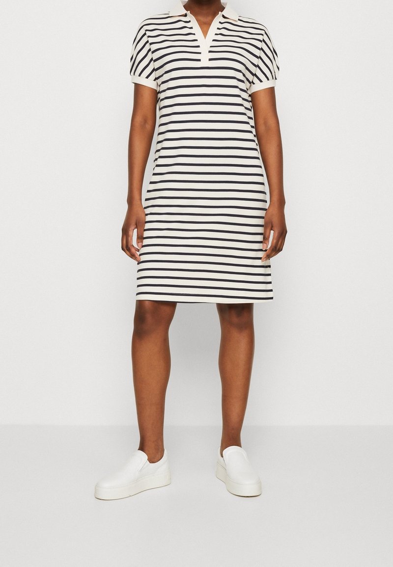 Femme portant une robe à manches courtes avec col, rayée horizontalement en noir et blanc, et des chaussures blanches sans lacets, debout devant un fond blanc.