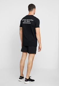 Black short-sleeve t-shirt, cotton material, features white text "National Aeronautics and Space Administration" on the back. Worn with black shorts.