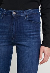 Primer plano de una persona que lleva jeans ajustados de color azul oscuro y una camiseta de manga larga negra, con una mano relajada a un lado.