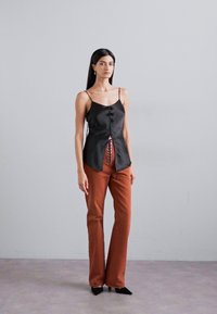 Sort satin camisole med frontknapper og splitdetalje, parret med rustfarvede tætsiddende bukser. Modellen har spidse sorte flade sko på.
