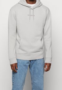 Sudadera con capucha gris claro con cordón, bolsillo canguro y logotipo bordado de "Hollister". Hecha de tela suave con puños y dobladillo acanalados.