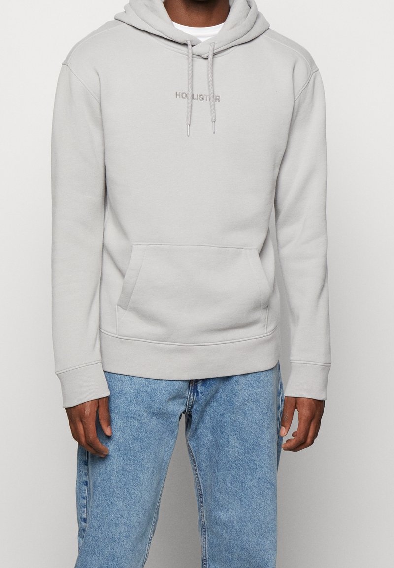 Sudadera con capucha gris claro con cordón, bolsillo canguro y logotipo bordado de "Hollister". Hecha de tela suave con puños y dobladillo acanalados.