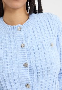 Ljusblå strukturstickad kavaj med ribbat mönster, prydd med kristall-liknande knappar och två framfickor. Mjukt material med en lätt lyster.
