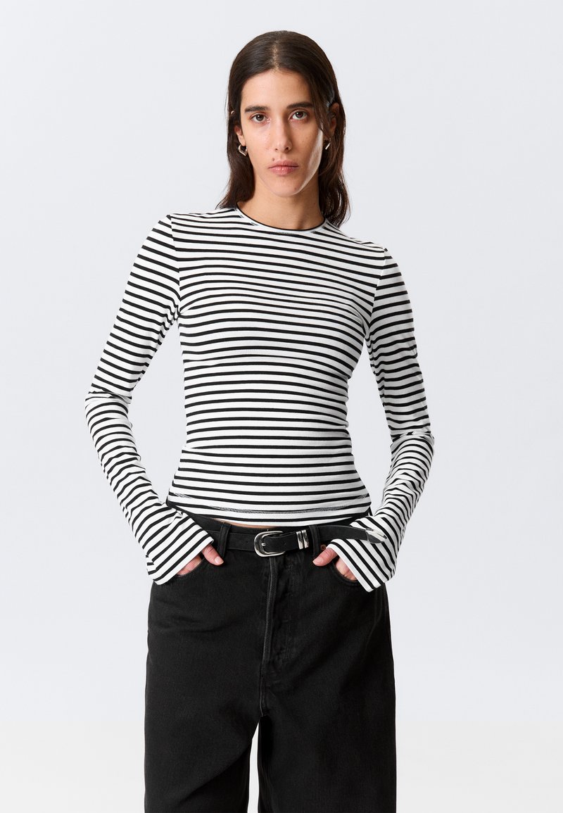 Sort og hvid stribet langærmet skjorte med tætsiddende pasform og udvidede manchetter, kombineret med højtaljede sorte bukser og et bælte.