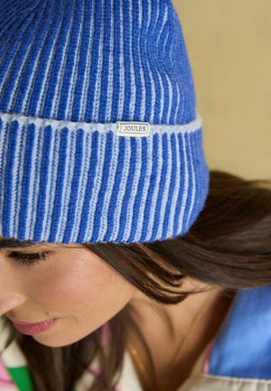 Žena u plavoj i bijeloj prugastoj pletenoj kapici s natpisom "Joules", s dugom smeđom kosom i ružičastim ružem za usne, vidljiva u mekom prirodnom svjetlu.